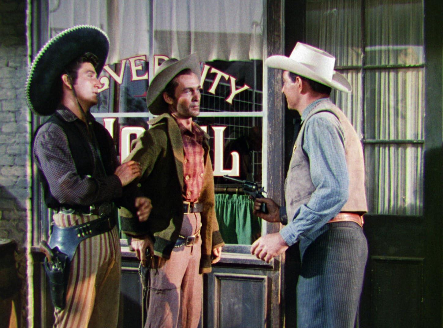 Drei Männer in Cowboy-Kleidung. Einer zielt mit einer Waffe. Einer wird von einem anderen gehalten. Im Hintergrund ist ein Laden zu sehen.