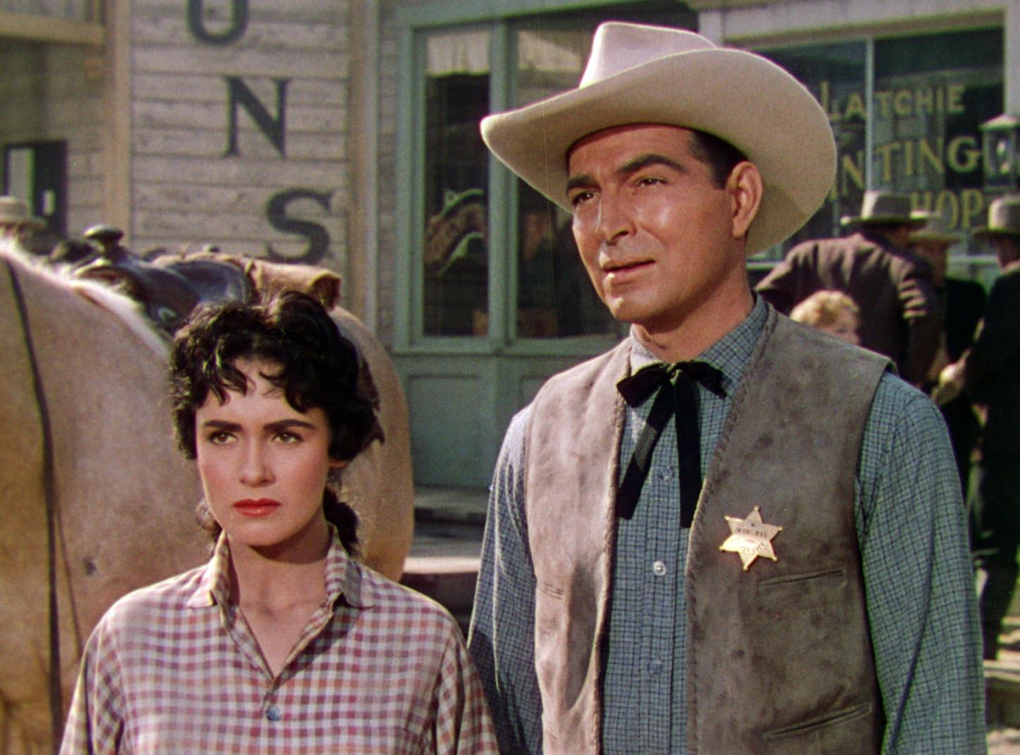 Ein Mann mit Cowboyhut und eine Frau in kariertem Hemd stehen vor einem Gebäude. Der Mann trägt ein Sternabzeichen.