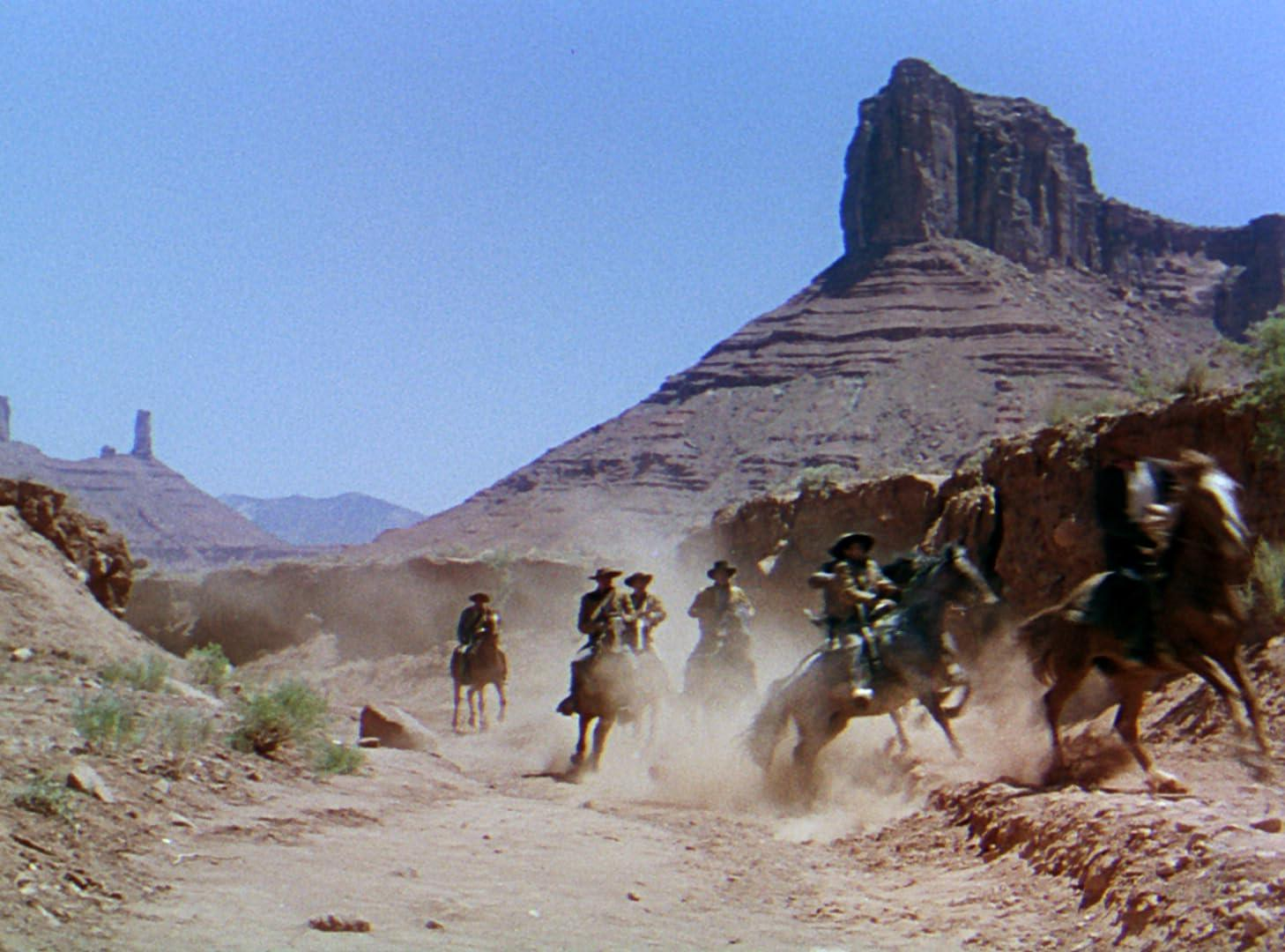 Cowboys auf Pferden reiten durch einen staubigen Canyon. Rote Felsformationen und ein klarer blauer Himmel bilden den Hintergrund.