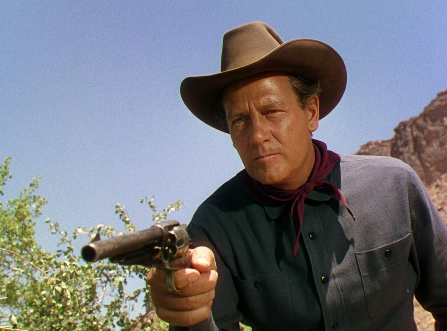 Ein Mann mit Cowboyhut zielt mit einer Waffe. Er trägt ein dunkles Hemd und einen roten Schal. Der Hintergrund zeigt eine Wüstenlandschaft.