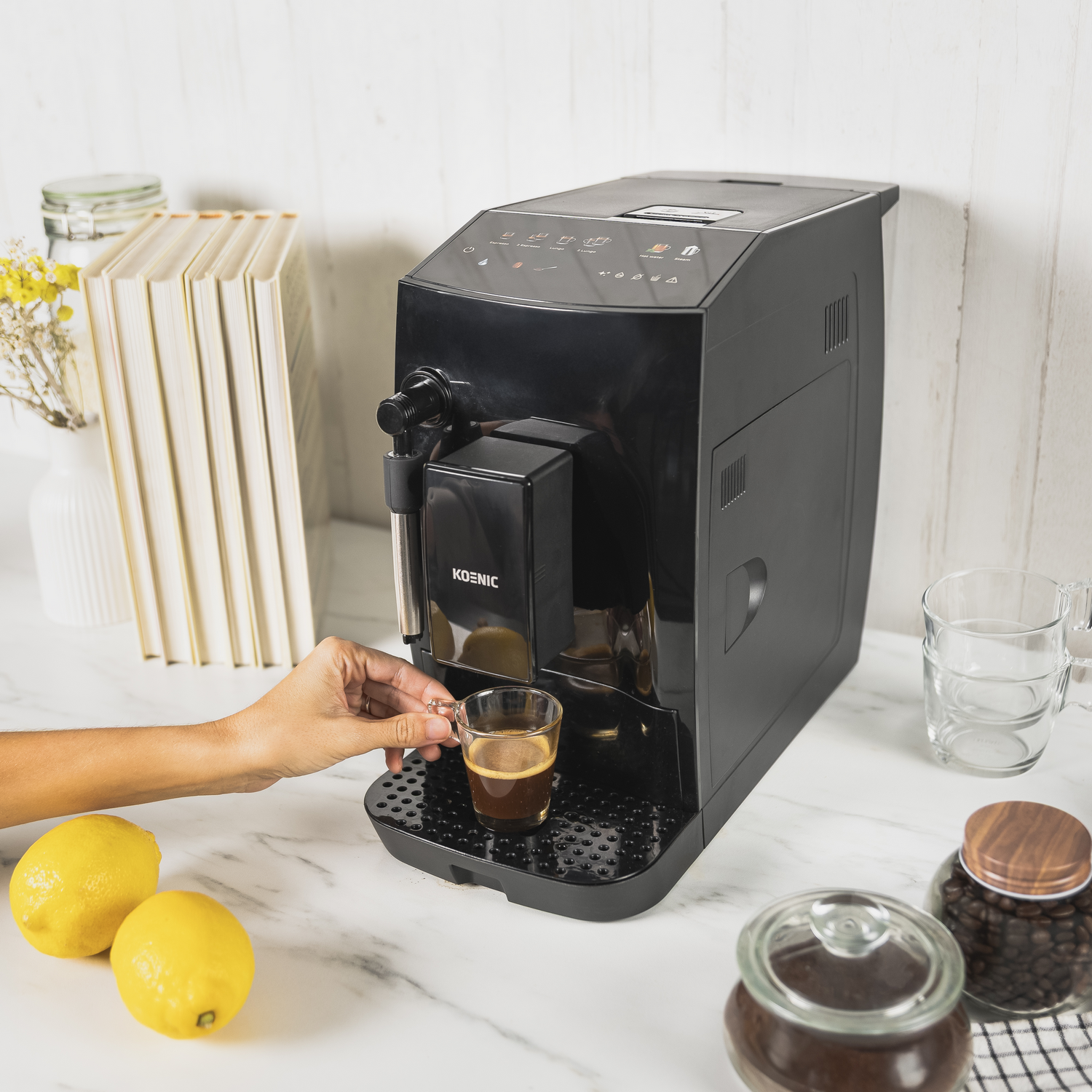 Una cafetera negra dispensa espresso en un vaso. Dos limones y frascos con granos de café están sobre una encimera de mármol.