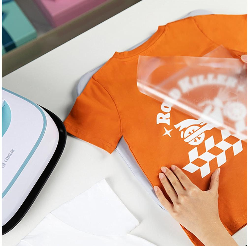 Ein orangefarbenes T-Shirt mit Design wird bedruckt. Eine Hand zieht das transparente Papier ab. Links ist ein weißes Bügeleisen.