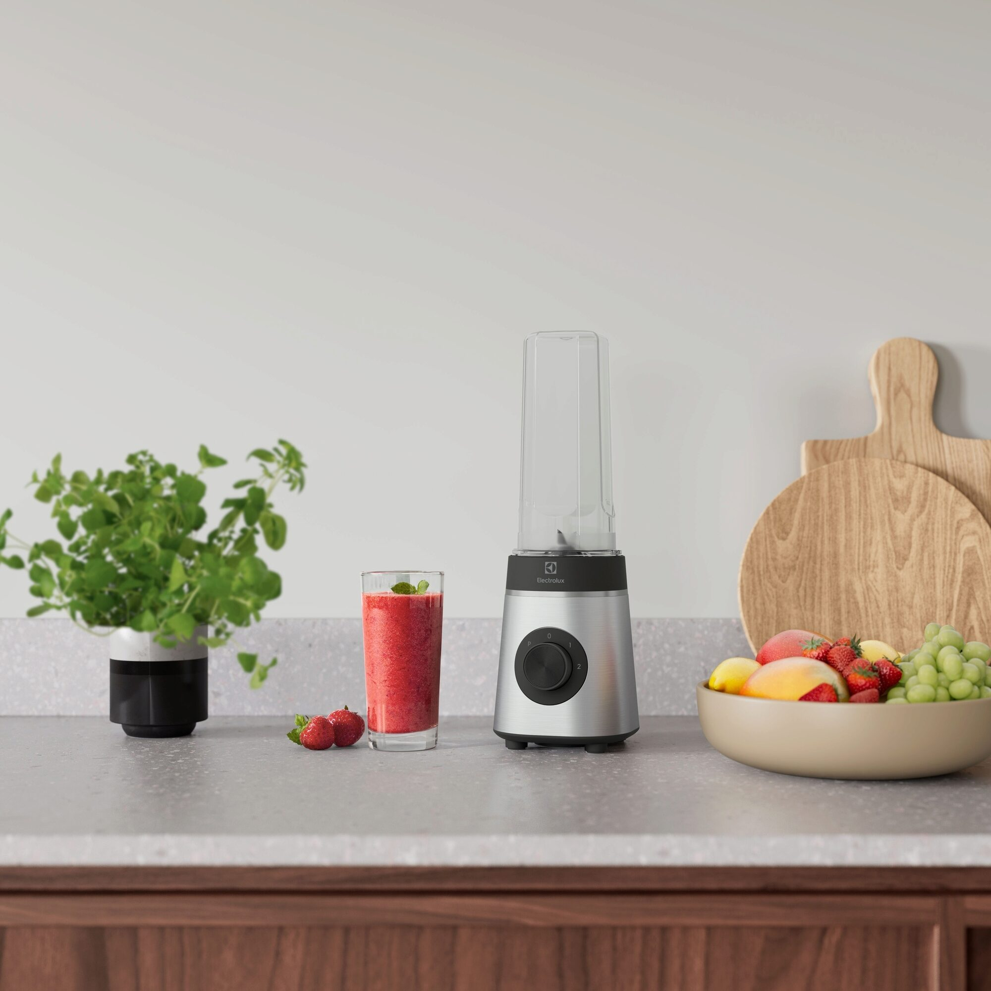 Bir blender, bir bardak smoothie, çilek ve bir kasede meyveler bir tezgahta duruyor. Bir bitki ve kesme tahtaları da mevcut.