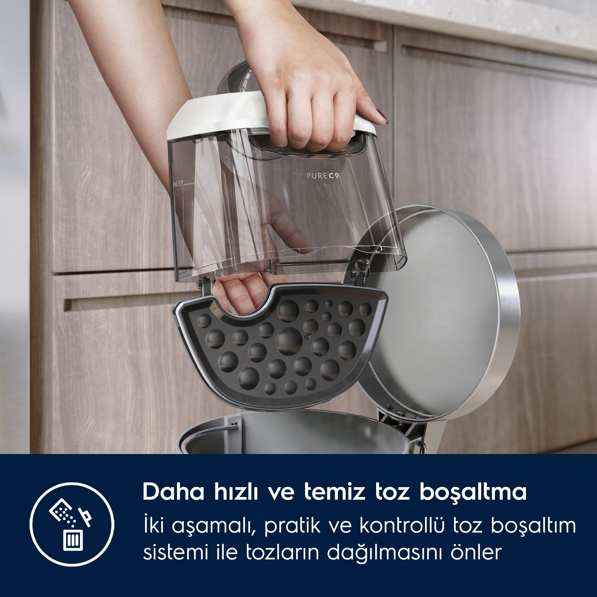 Bir kişi bir elektrik süpürgesinin toz kabını açık tutuyor. Şeffaf ve siyah bir filtreye sahip. Arka plan ahşap damarlı dolaplara sahip.