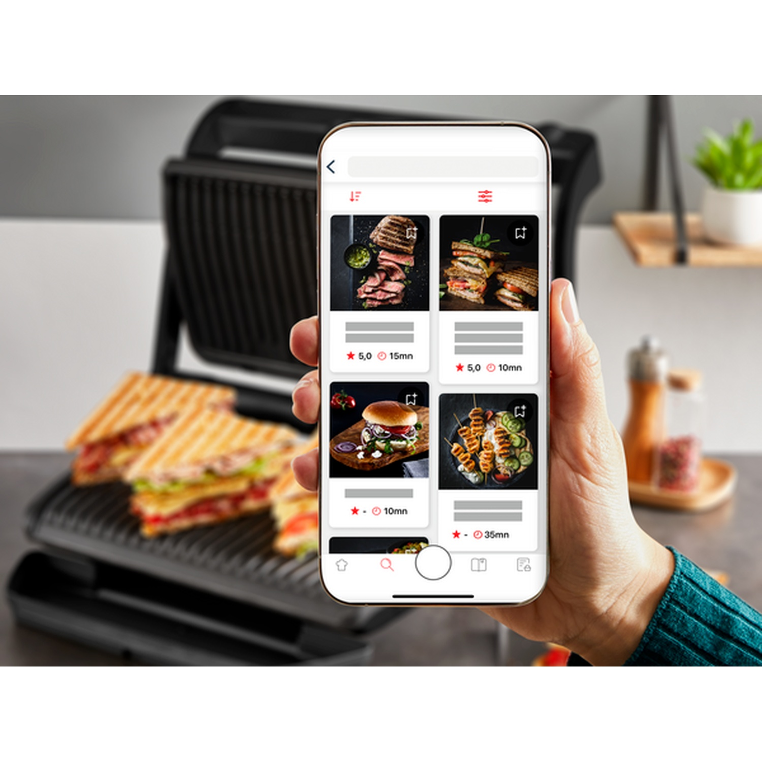 Une main tenant un téléphone affichant des images de nourriture. Un grill à sandwichs et des sandwichs en arrière-plan.