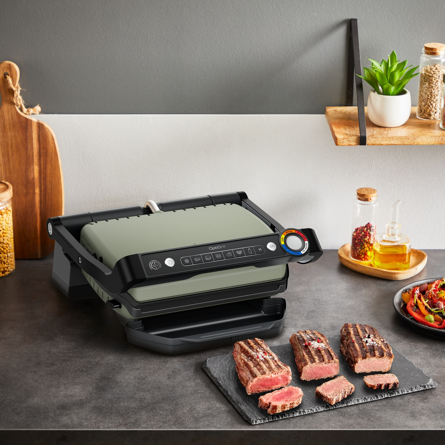Gril électrique avec steaks cuits sur une ardoise. Une planche en bois, des épices et une plante sont également sur le comptoir.