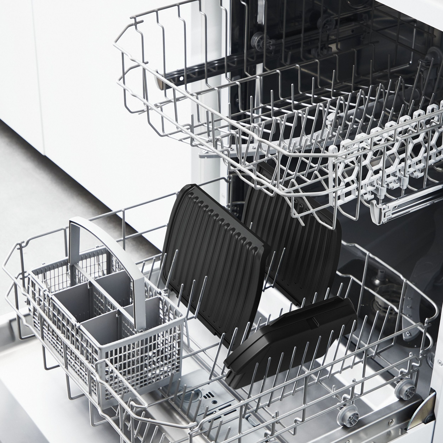 Intérieur de lave-vaisselle avec plaques de grillades noires et un panier à ustensiles. Des supports argentés maintiennent les objets. La porte du lave-vaisselle est ouverte.