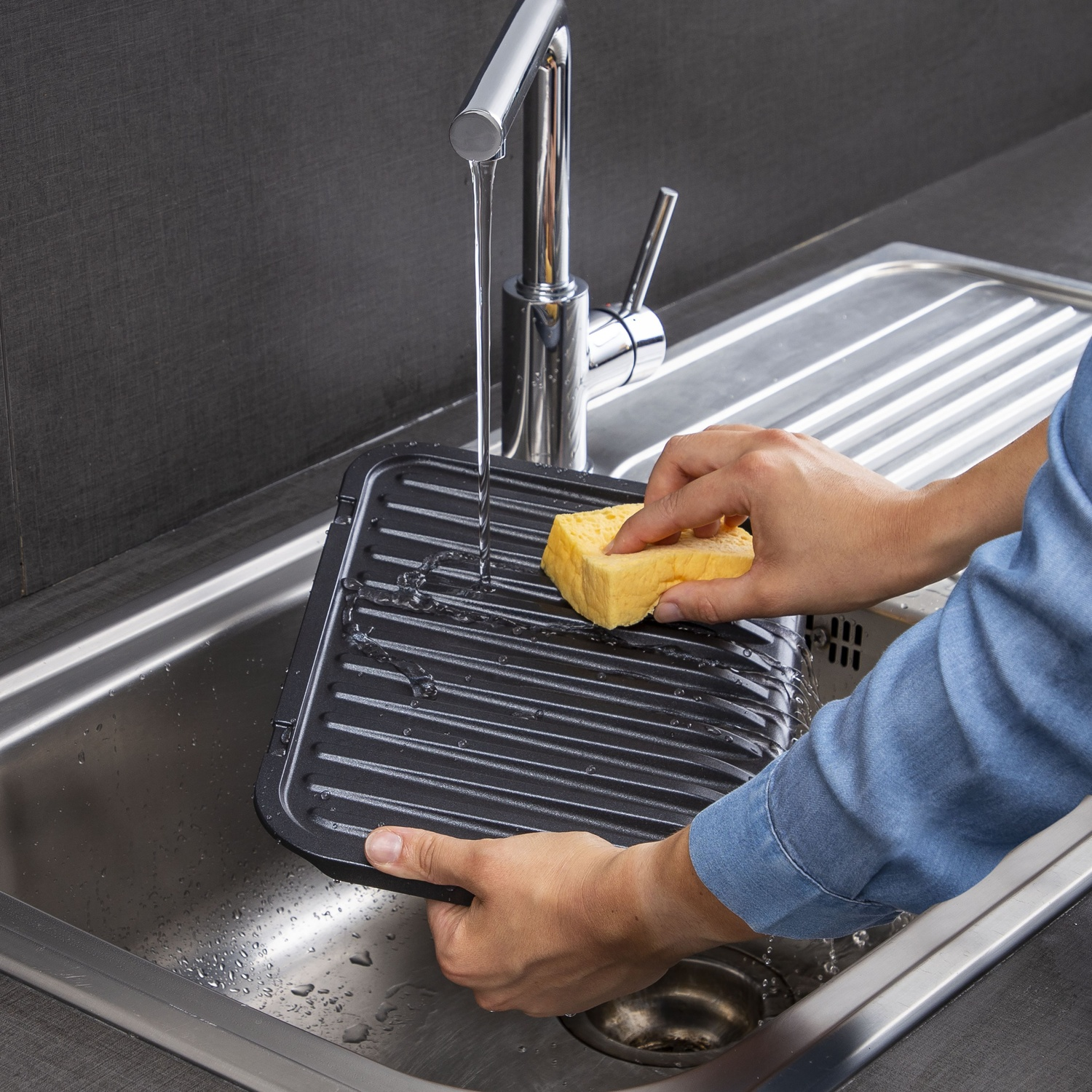 Une personne lave une plaque de cuisson noire avec une éponge jaune dans un évier en acier inoxydable sous l'eau courante.