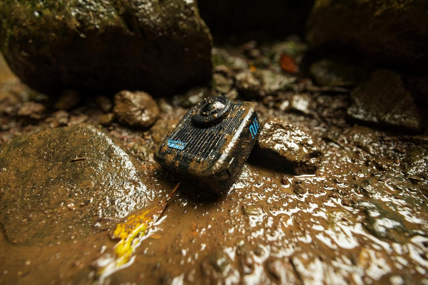 Una cámara GoPro negra cubierta de barro descansa en agua fangosa entre rocas. La lente de la cámara mira hacia arriba.