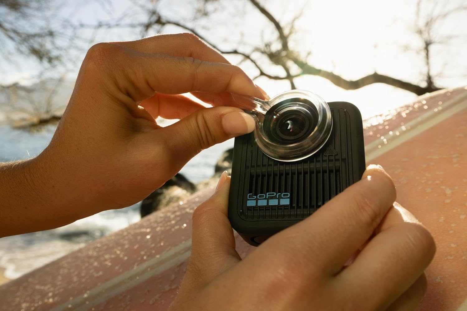 Manos sosteniendo una cámara GoPro con un accesorio de lente. La cámara está sobre una tabla de surf, con una escena de playa borrosa al fondo.