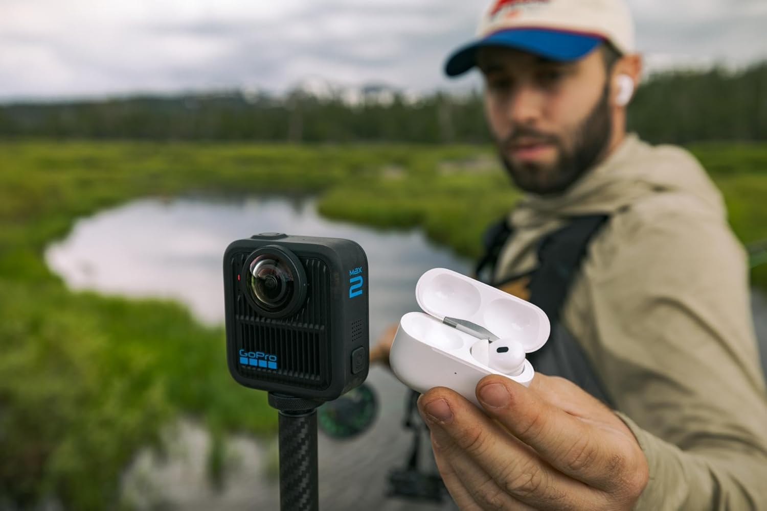Hombre sosteniendo estuche de AirPods, cámara GoPro Max 2 en trípode y paisaje verde con río al fondo.