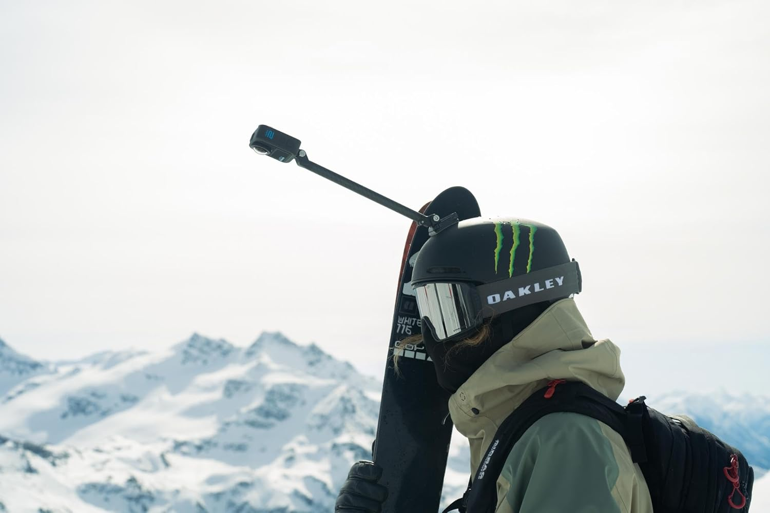 Esquiador con casco y gafas, sosteniendo una tabla de snowboard. Una cámara está sujeta al casco. Montañas nevadas al fondo.