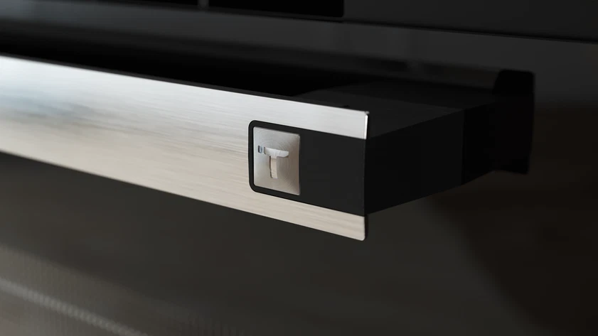 Primer plano de un tirador de cajón de metal cepillado con marco negro. Se ve un botón plateado con forma de 'T'.
