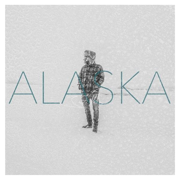 Ein Mann in einem karierten Hemd steht im Schnee. Das Wort 'ALASKA' steht über dem Bild. Der Hintergrund ist eine verschneite Landschaft.