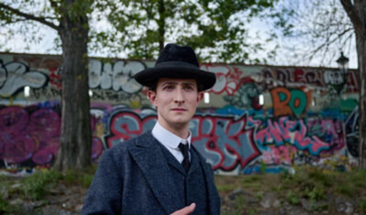 Ein Mann mit schwarzem Hut und Anzug steht vor einer Graffiti-Wand. Bäume umrahmen die Szene. Die Wand hat farbenfrohe Kunst.