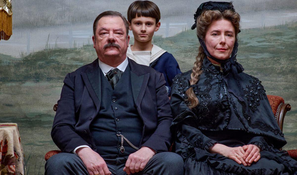 Ein Familienporträt. Ein Mann mit Schnurrbart und eine Frau in Schwarz sitzen mit einem Jungen im Hintergrund. Der Hintergrund ist ein Landschaftsbild.