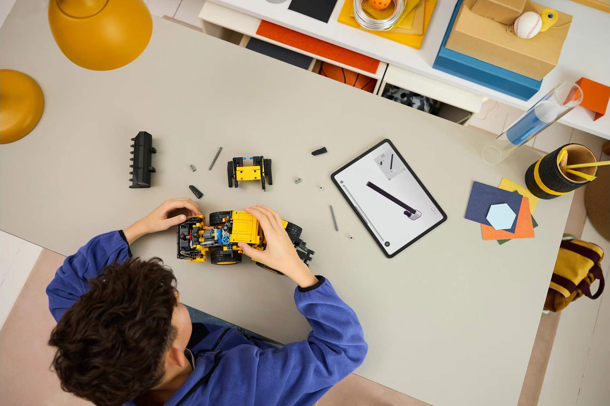 Une personne assemble un camion jouet jaune sur une table, avec une tablette affichant des instructions et divers autres objets.
