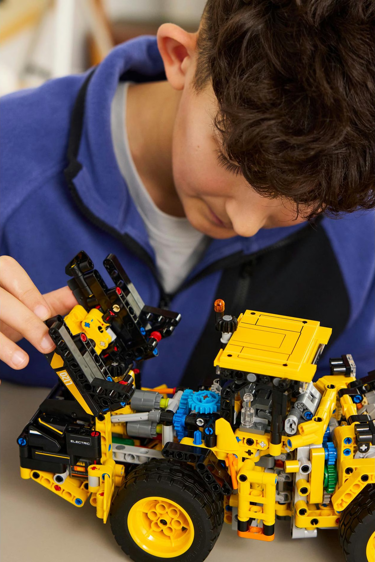 Un garçon assemble un modèle Lego Technic jaune et noir. Le modèle a des roues et des engrenages. Le garçon sourit.