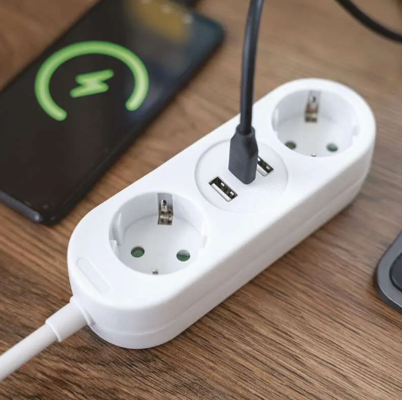Egy fehér elosztó USB portokkal és aljzatokkal egy fa felületen. Egy telefon töltődik zöld villám ikonnal.