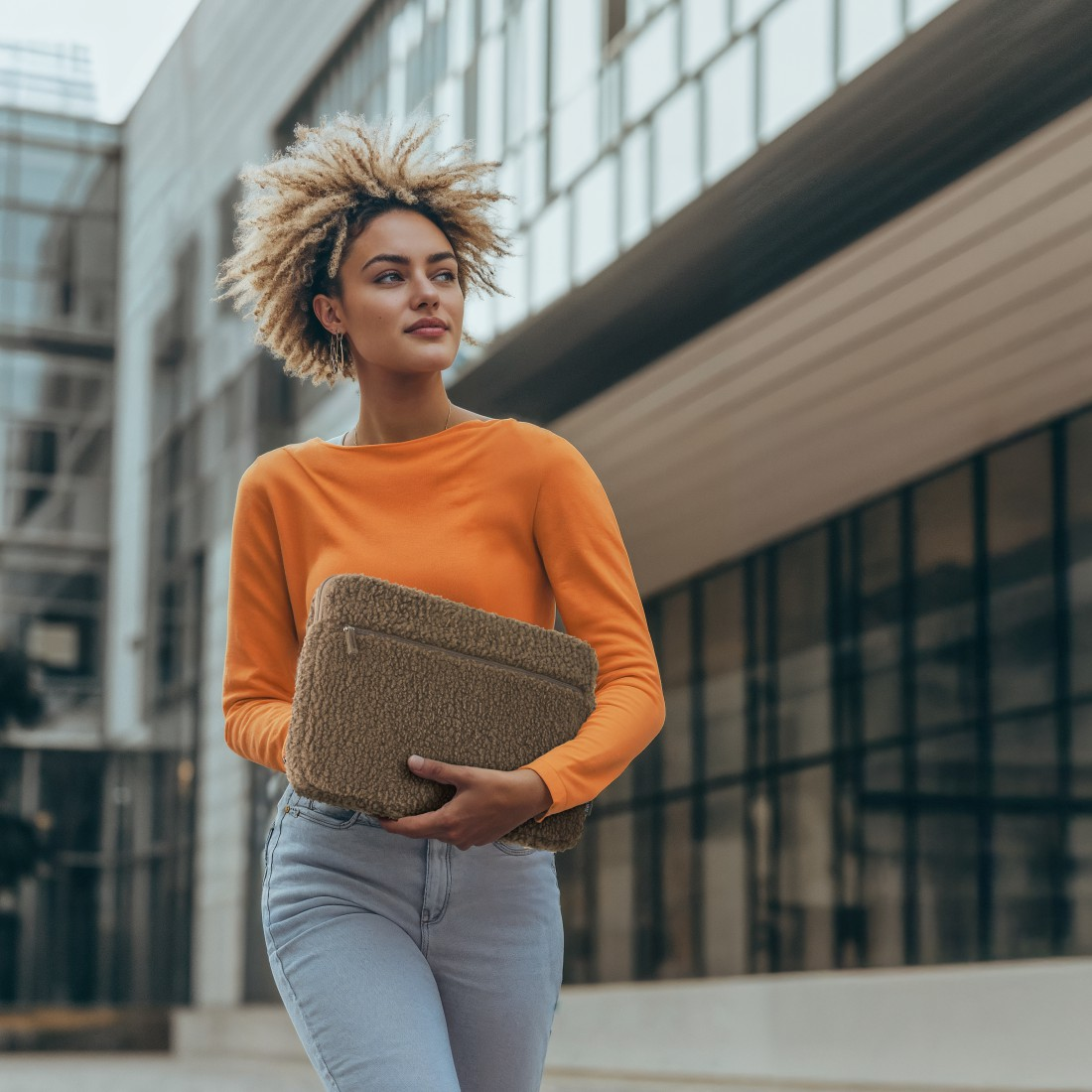 Frau mit lockigem Haar in orangefarbenem Oberteil und Jeans, hält eine Laptoptasche, geht vor einem modernen Gebäude.