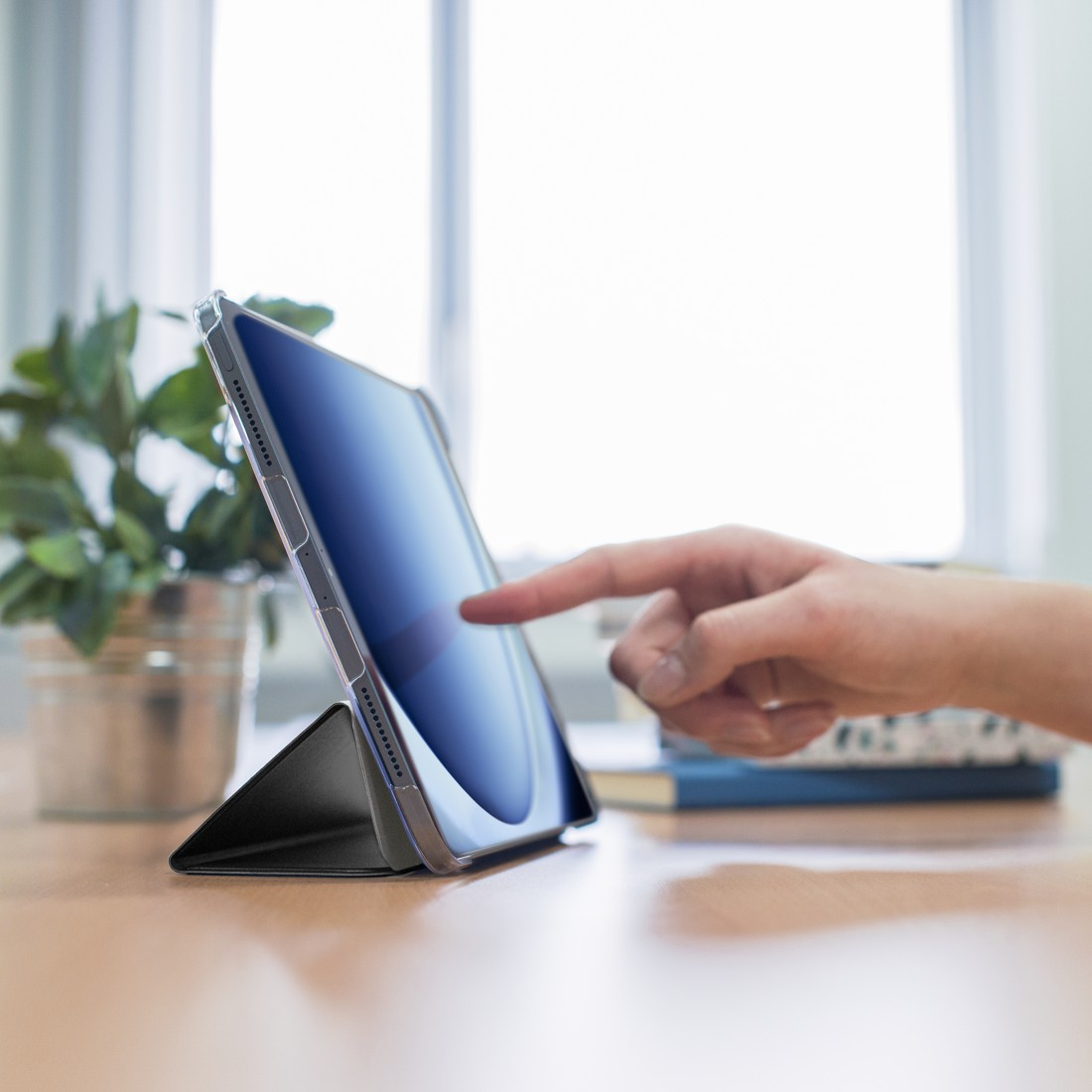 Eine Hand berührt einen Tablet-Bildschirm. Das Tablet befindet sich in einer Hülle und auf einem Holztisch. Eine Pflanze und Bücher sind im Hintergrund.