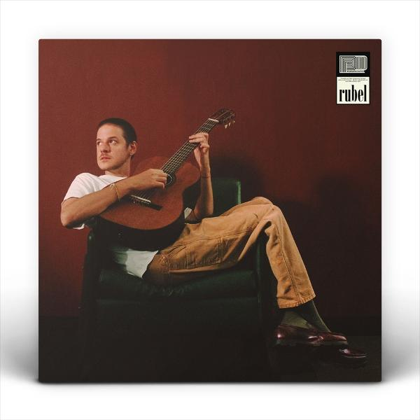 Ein Mann sitzt in einem Stuhl und spielt Gitarre. Er trägt ein weißes Hemd und braune Hose. Der Hintergrund ist eine dunkelrote Wand.