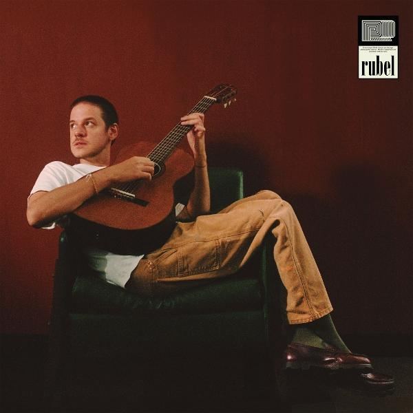 Ein Mann sitzt und spielt Gitarre, trägt ein weißes Hemd und braune Hose. Er sitzt in einem grünen Stuhl, mit dunkelrotem Hintergrund.