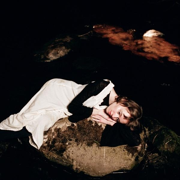 Eine Frau ruht auf einem bemoosten Felsen in der Nähe von Wasser. Sie trägt ein weißes Kleid und ein schwarzes Oberteil. Der Hintergrund ist dunkel.