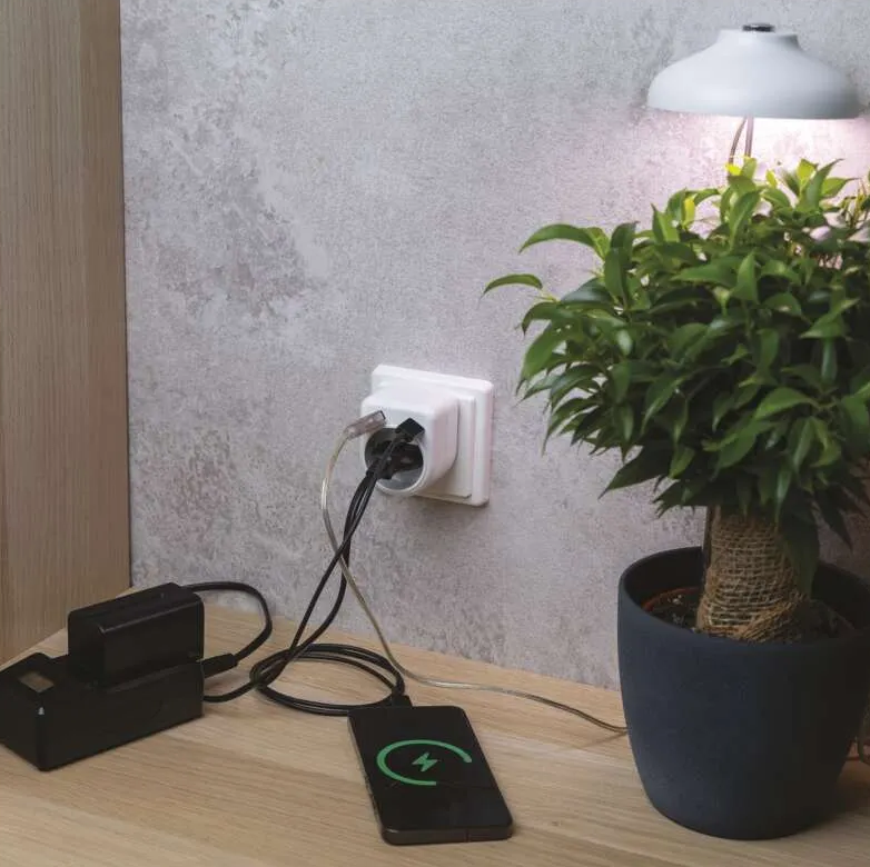 Egy telefon töltődik, egy fekete töltő és egy növény mellett. A telefon egy töltési ikont mutat. Egy elektromos aljzat látható.
