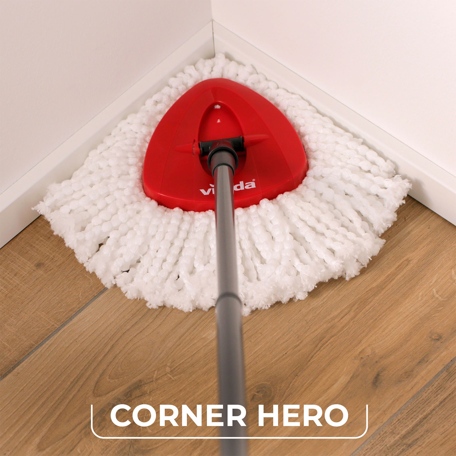 Una testa di mop rossa con fili bianchi è in un angolo. Il pavimento è in legno. Il muro è bianco. Il mop dice 'vida'.