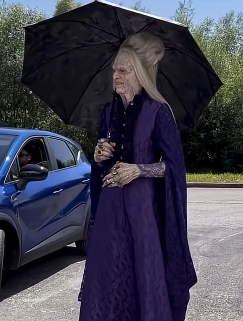 Eine ältere Person in lila Kleid und mit einem Regenschirm steht in der Nähe eines blauen Autos. Die Person hat blasse Haut und trägt Ringe.