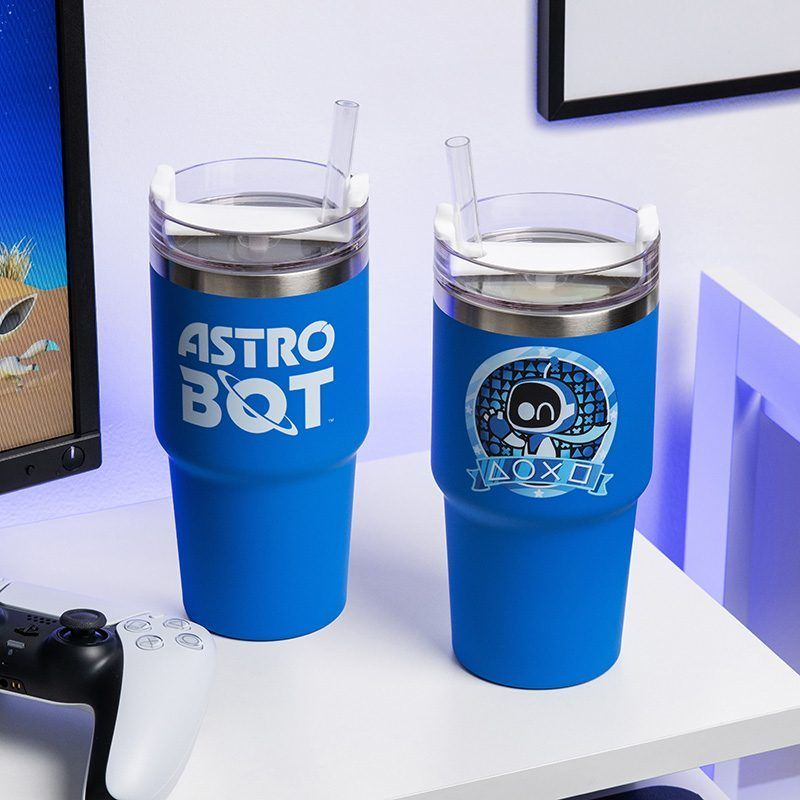 Két kék Astro Bot pohár szívószállal egy fehér felületen, egy kontroller és egy monitor mellett.
