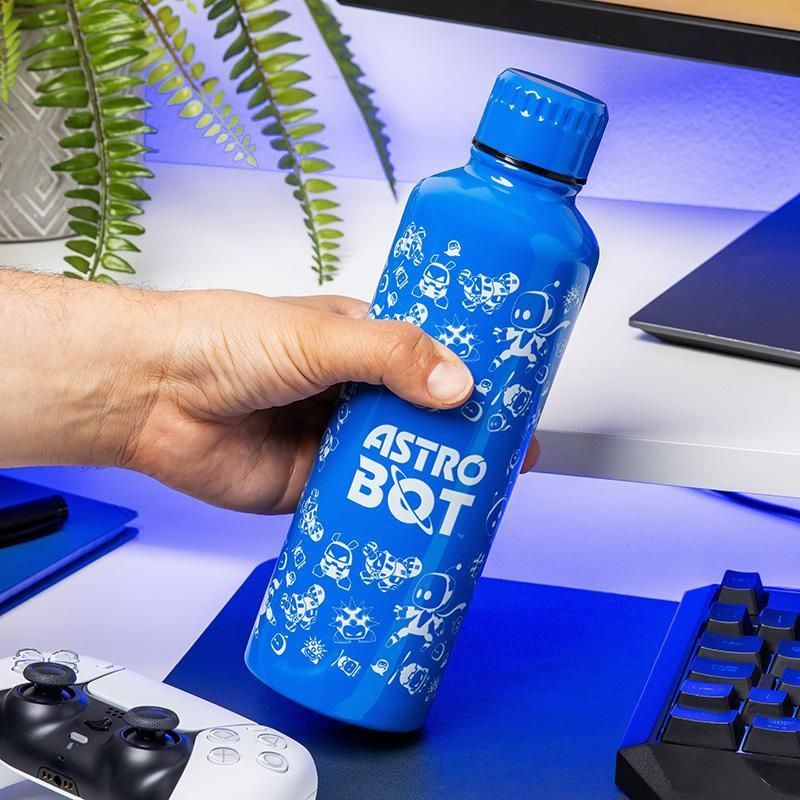 Egy kéz egy kék Astro Bot vizespalackot tart. A palackon fehér rajzfilm minták vannak. Egy PlayStation vezérlő és egy billentyűzet látható.