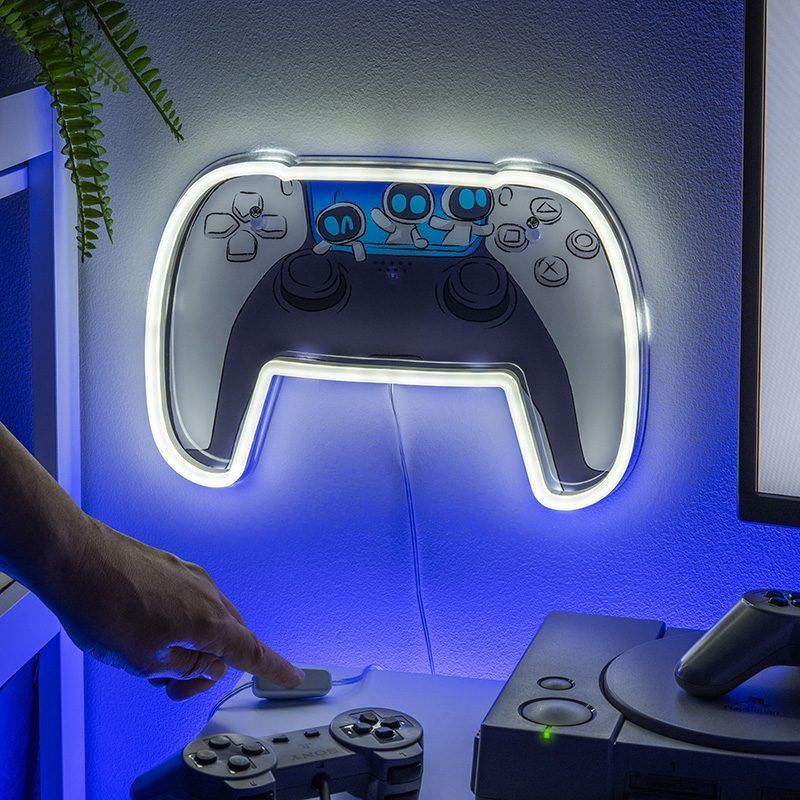 Egy világító PlayStation-kontroller alakú fali lámpa. Egy kéz éppen egy gombot nyom meg egy asztalon, egy PS1 és egy kontroller mellett.