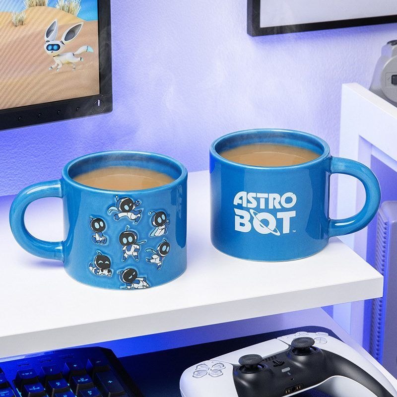 Két kék bögre kávéval. Az egyik Astro Bot karakterekkel, a másik 'Astro Bot' felirattal. A háttérben egy játékkal rendelkező képernyő látható.