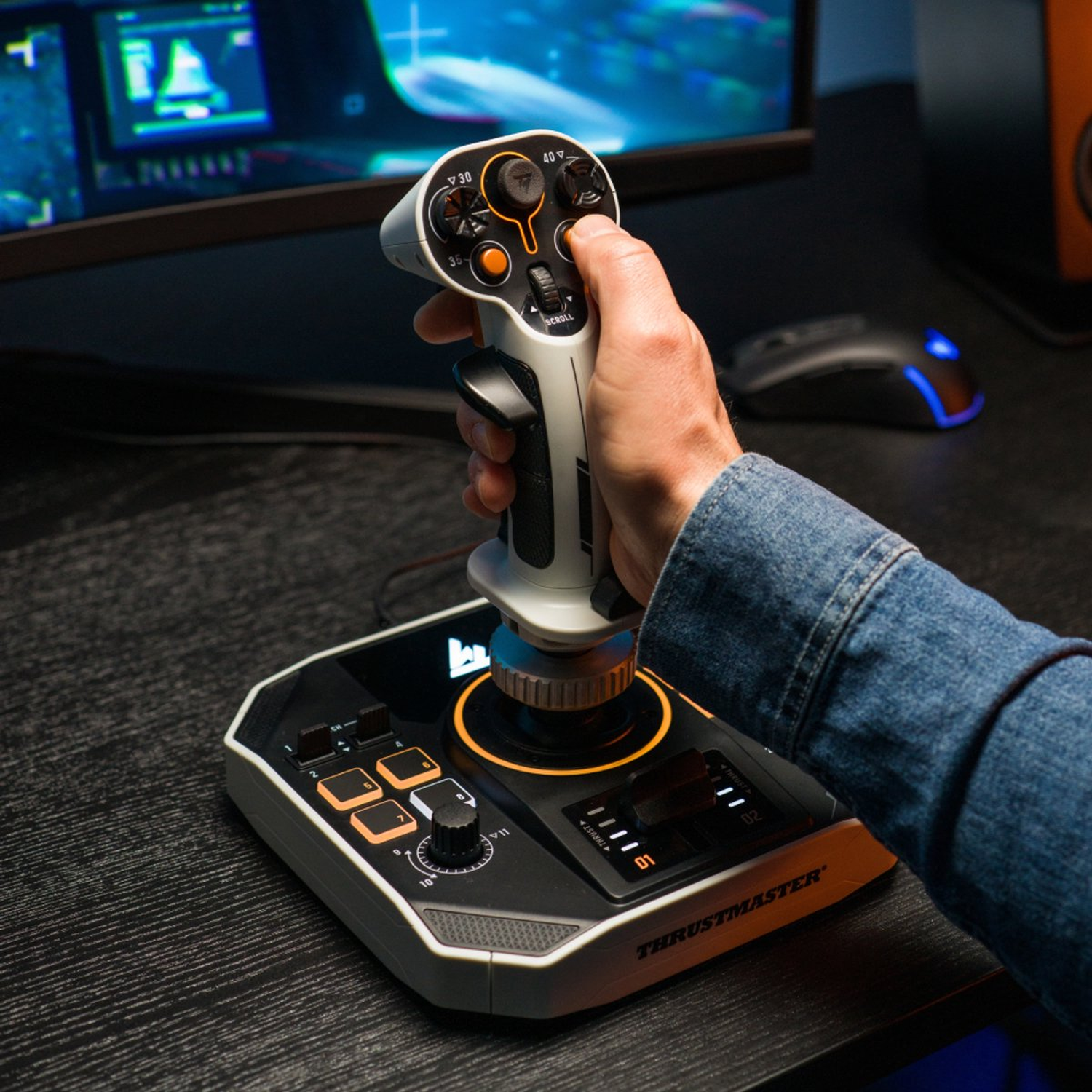 A person is holding a white and black joystick with orange details. It is sitting on a black table with a computer monitor in the background.