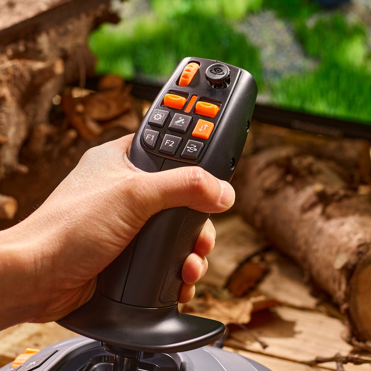 Een hand die een zwarte controller met oranje knoppen vasthoudt. De achtergrond toont hout en een scherm met groen gras.