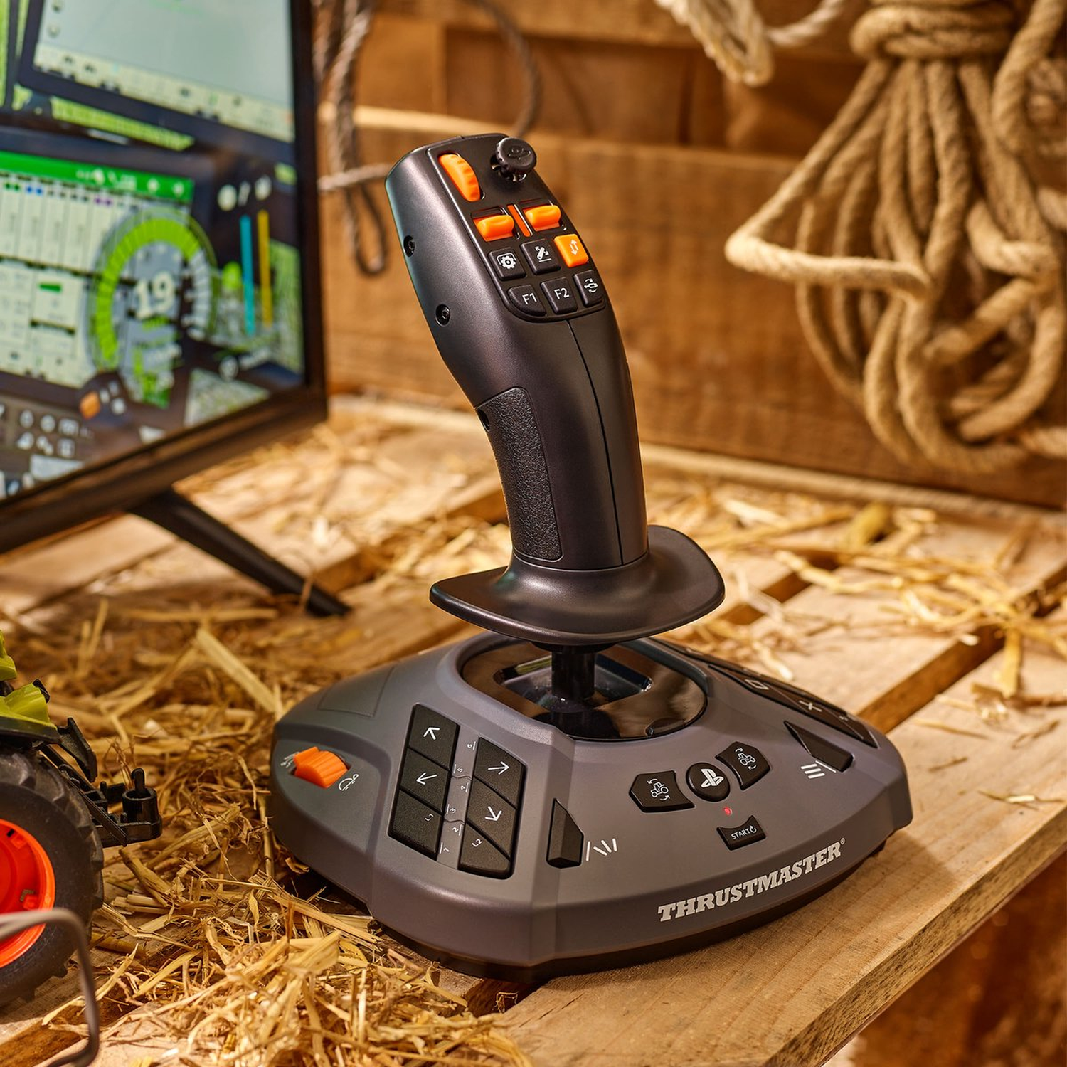 Een Thrustmaster joystick staat op hout, omringd door hooi. Een scherm toont een boerderijspel. Een touw en een speelgoedtrekker zijn ook in de scène.
