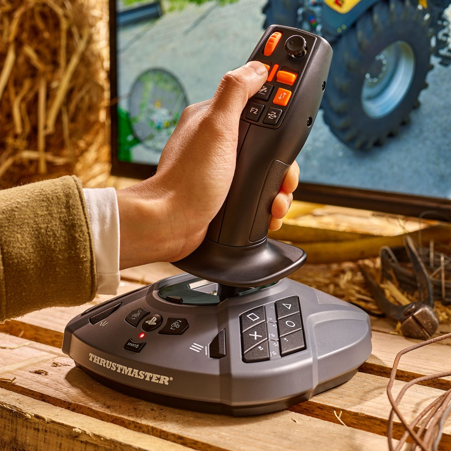 Een hand die een zwarte Thrustmaster joystick vasthoudt met oranje knoppen, voor een scherm met een tractor.