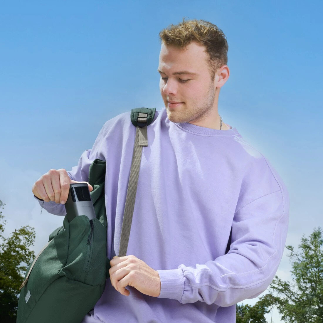 Mężczyzna w fioletowym swetrze wkłada przedmiot do zielonego plecaka. Jest na zewnątrz z drzewami i błękitnym niebem w tle.