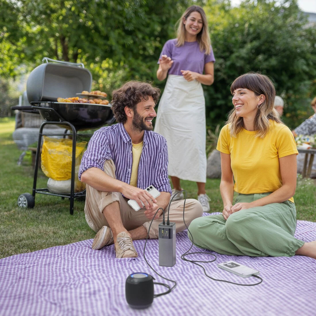 Trzy osoby na pikniku. Dwie siedzą i rozmawiają, jedna stoi. Na trawniku widoczny jest również grill i głośnik.