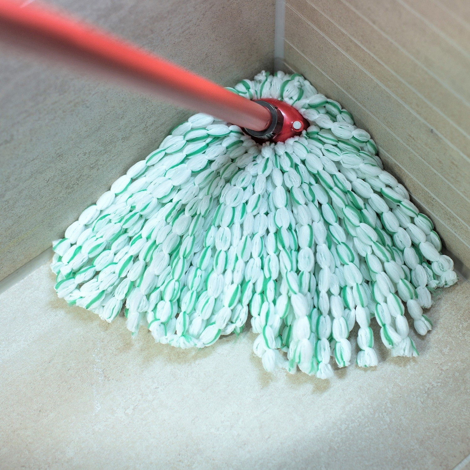 Un mocio con manico rosso e testina bianca e verde è posizionato su un pavimento piastrellato in un angolo. Le pareti sono beige.