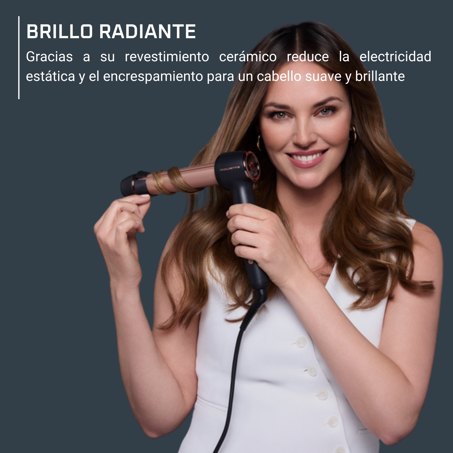 Mujer sonríe, riza el cabello con un rizador negro y oro rosa. Top blanco. Fondo oscuro.