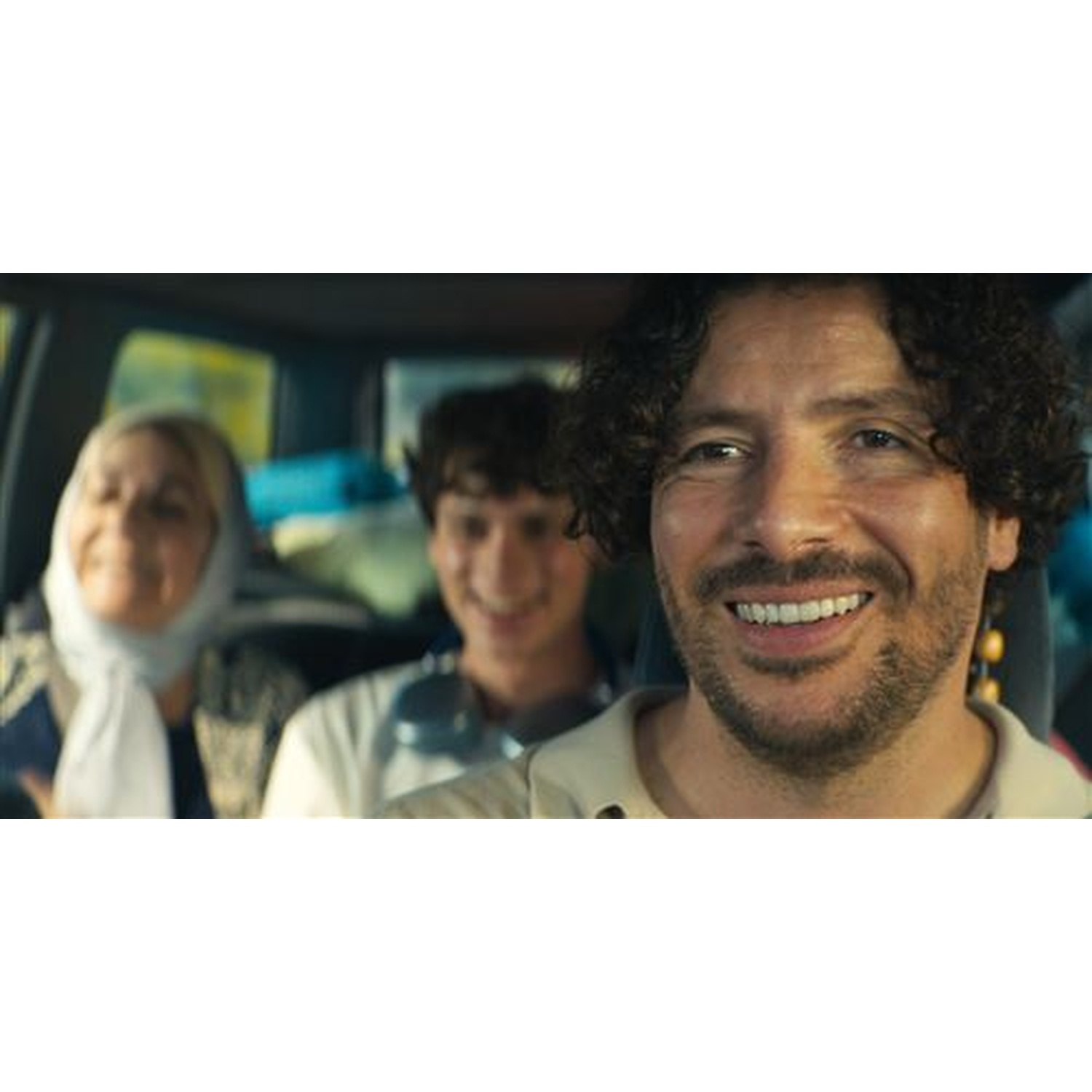 Trois personnes dans une voiture. Un homme souriant aux cheveux bouclés devant, et deux autres personnes derrière.