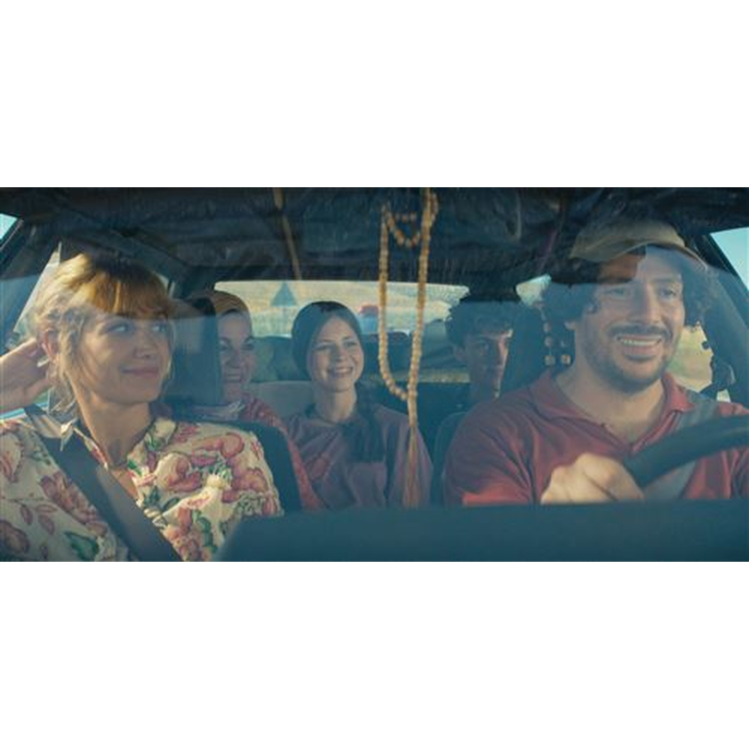 À l'intérieur d'une voiture, on voit cinq personnes. Le conducteur sourit. Les passagers à l'arrière sourient. Le soleil brille à travers le pare-brise.