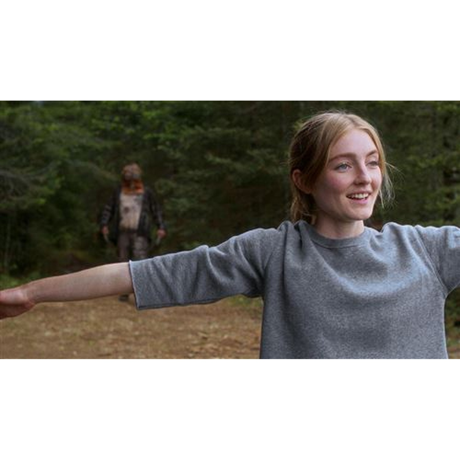 A young woman in a gray sweater smiles, arms outstretched. A man in the background stands in the woods.