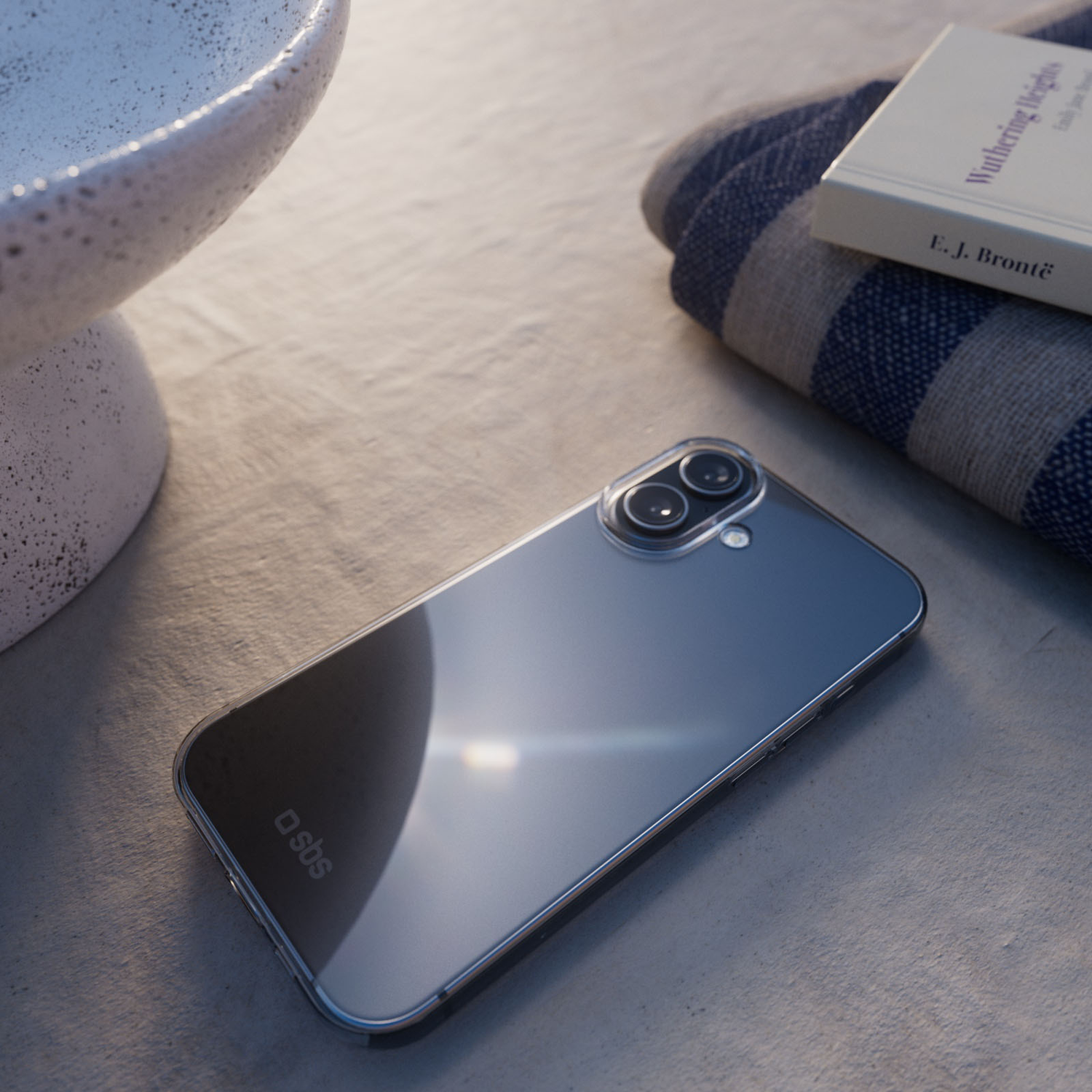 A phone with a clear case lies on a table, next to a book and a striped towel. A white bowl is also visible.