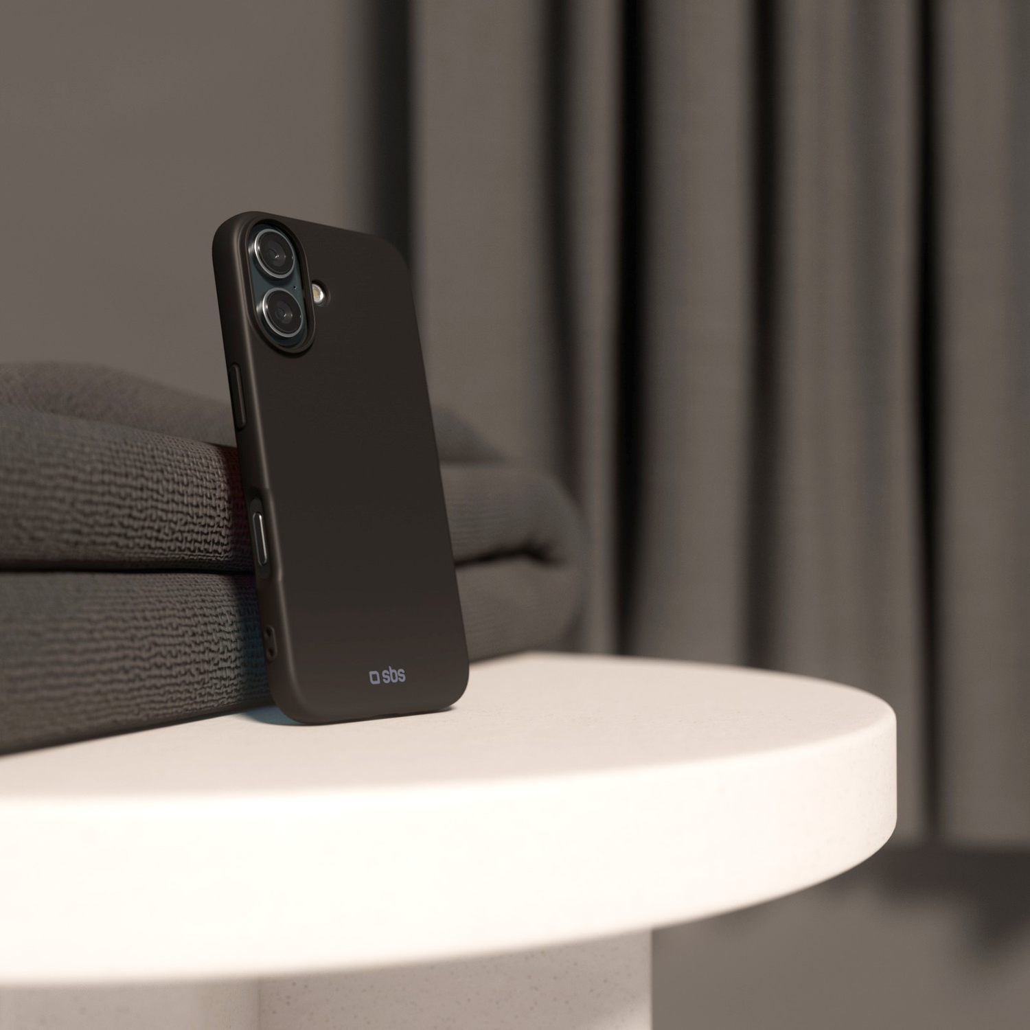 Une coque de téléphone noire est posée sur une table blanche à côté de serviettes grises pliées. Un rideau sombre en arrière-plan.
