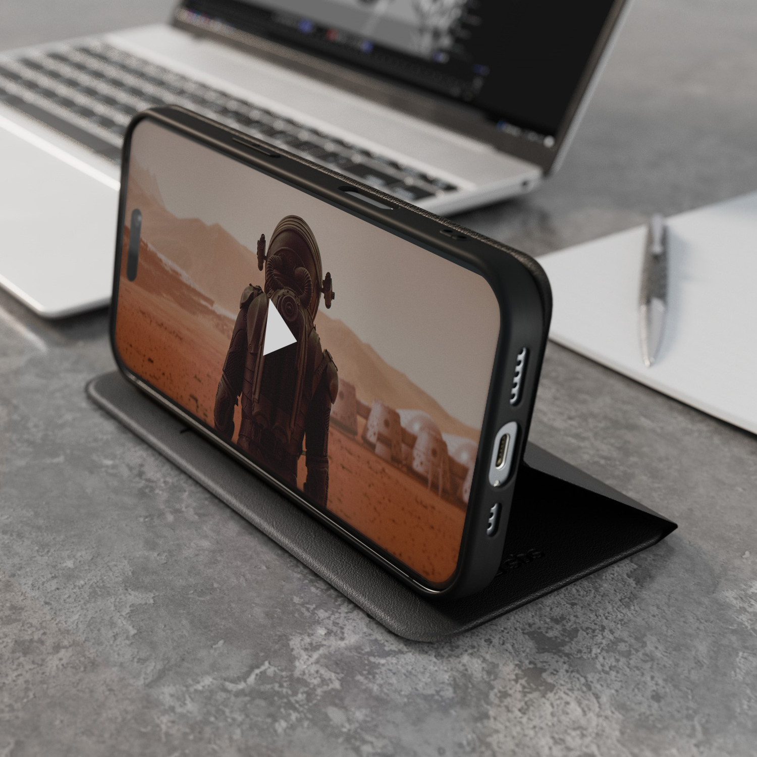 Telefoon die een video op een standaard afspeelt. Laptop, pen en notitieblok op de achtergrond, alles op een getextureerd grijs oppervlak.