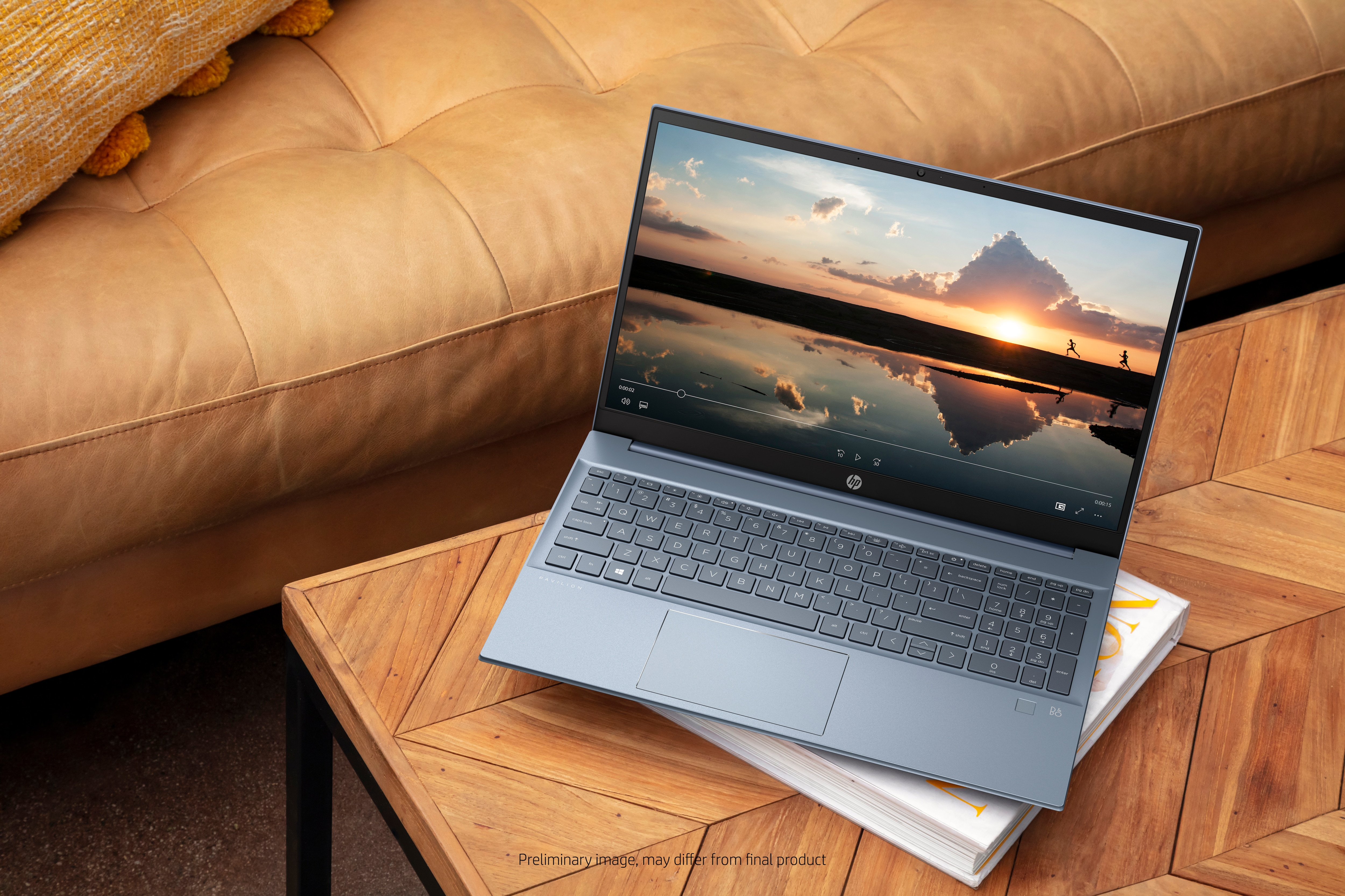 Laptop na drewnianym stole, wyświetlający wideo z zachodem słońca. Obok sofy z żółtą poduszką.